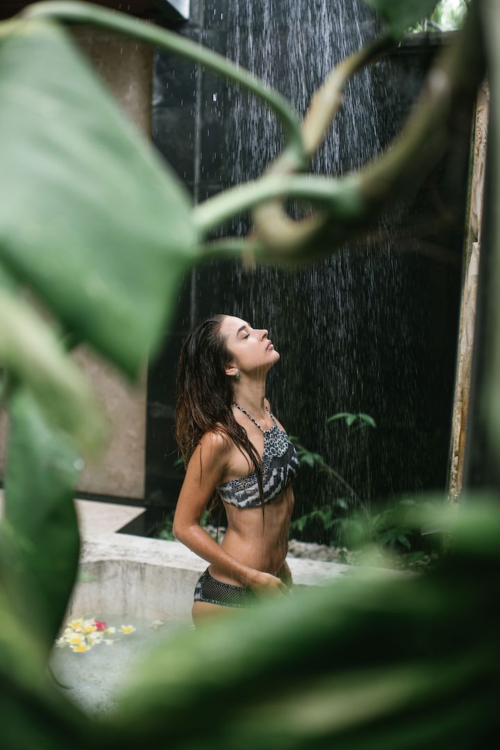 A young woman refreshing her body by closing her eyes too the shower in a shower pool with ambiance type flowers in the pool.