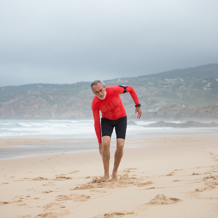 Man in Red Shirt and Black Shorts Feeling Pain in His Legs during a walk in the beach wearing a fitness device in his left arm.