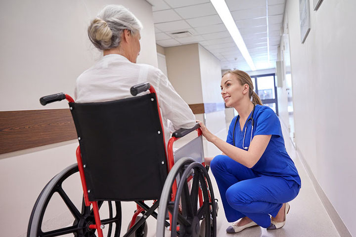 A female multiple sclerosis patient on a wheelchair talking to a nurse who is crouching down to her level.