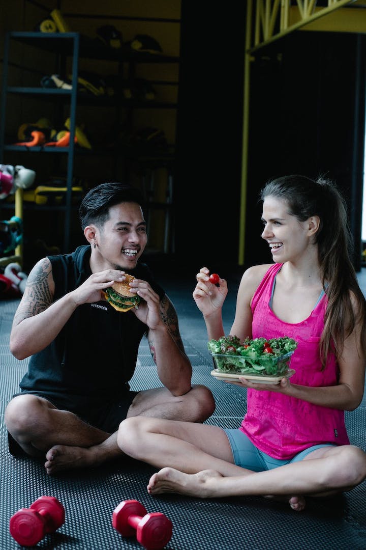 A man and a woman smiling and sitting as they hold different food items both with spinach to eat in a training environment.