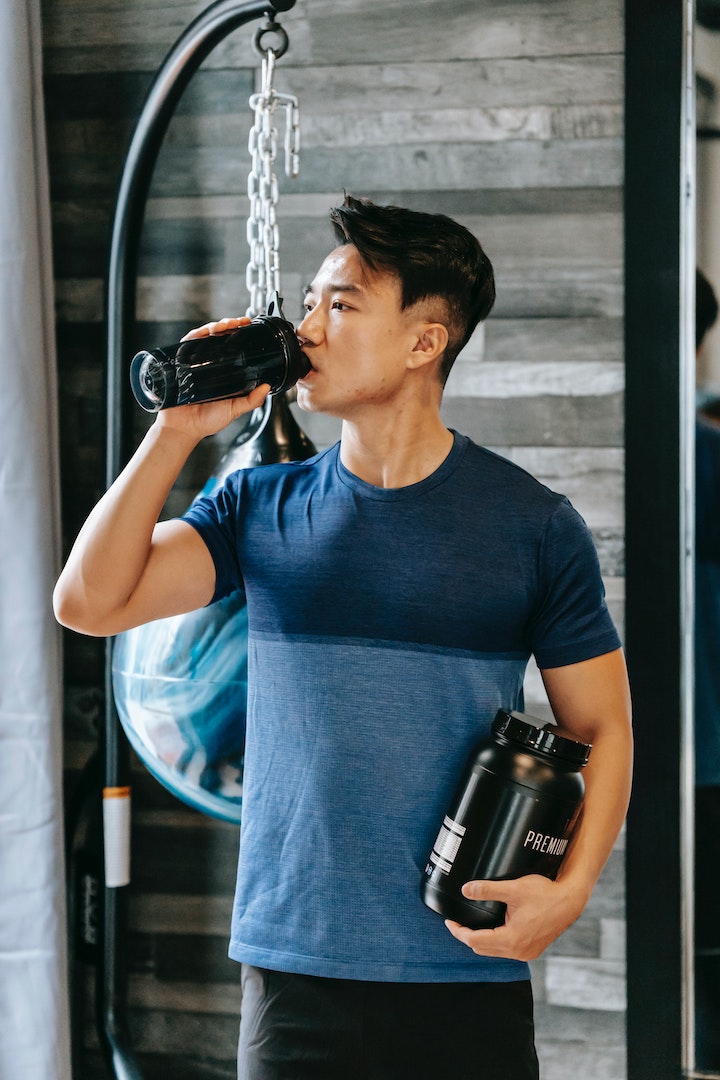 An Asian man drinking water from a bottle while holding a jar of sports nutritional supplement.