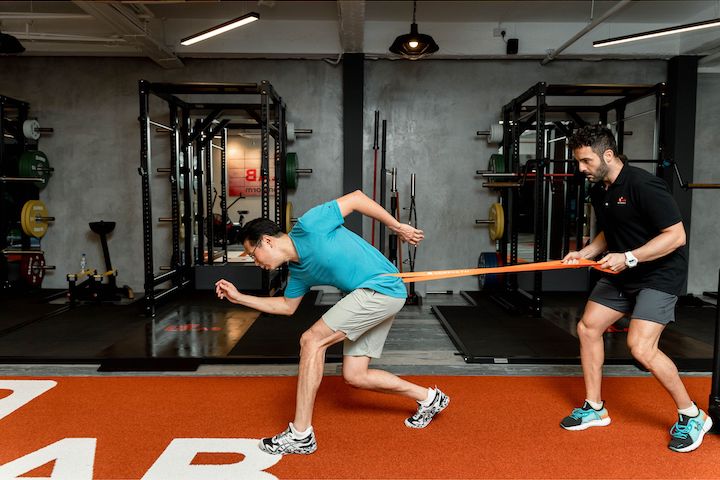 A personal trainer training an Asian man as if he's running as he pulls on an elastic band on tied to the trainee's waist.