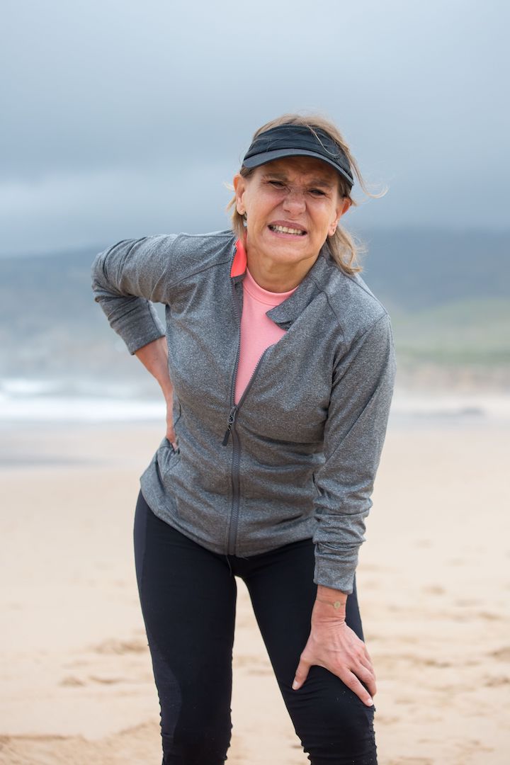 An middle-aged woman in gray zip-up jacket and black leggings standing on beach with one hand on her waist back and the other on her knee.