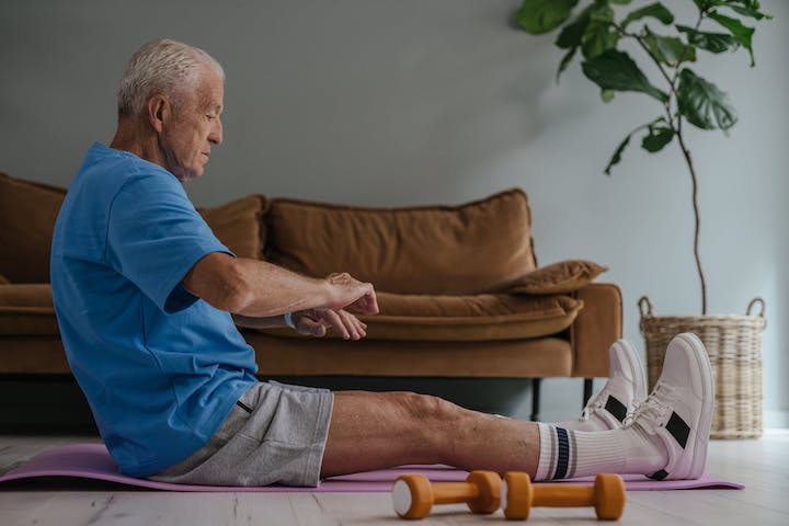 An elderly man during a break of his physical therapy session with wooden light-weight dumbbells beside his legs.