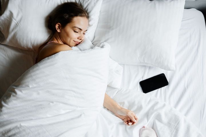 A woman sleeping under a blanket with a smile with a phone beside her.