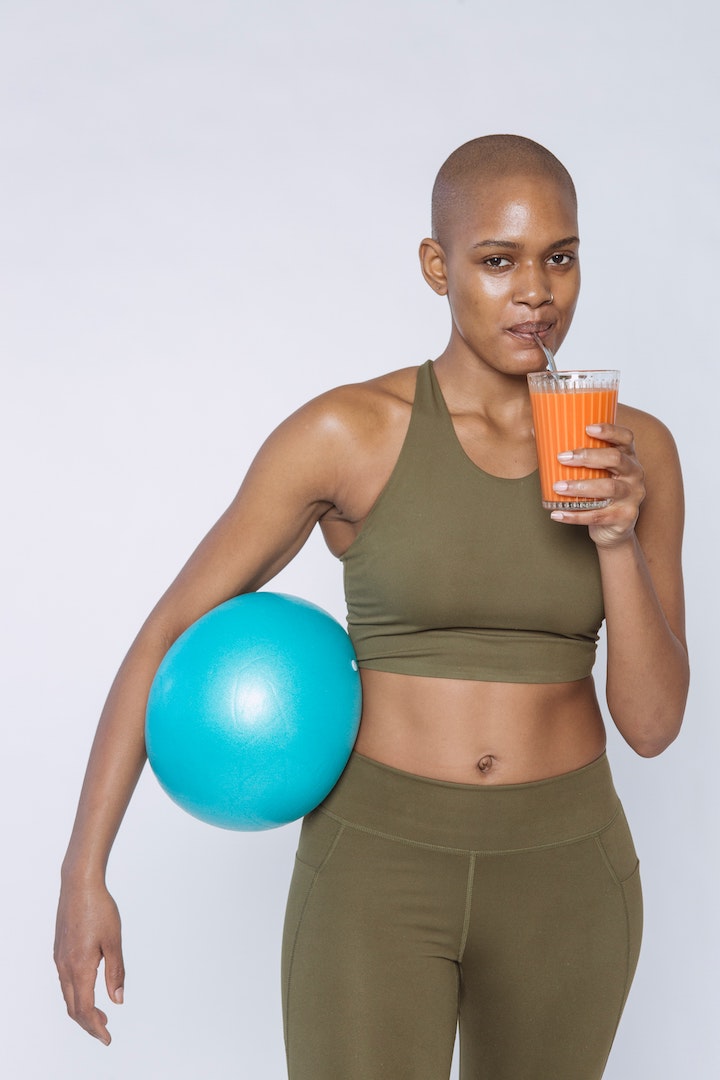 A bald ethnic woman drinking a carrot drink while holding a blue training ball while wearing training gear.