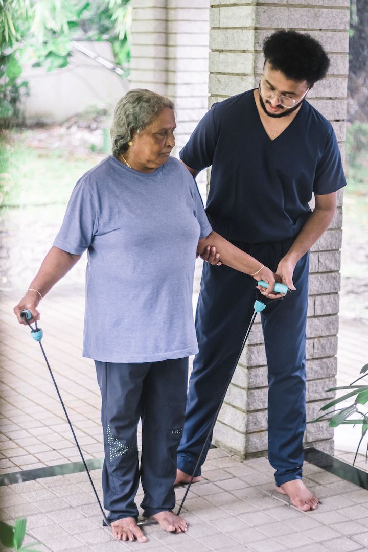 A male nurse helping an old woman exercise with a resistance band pressed under her feet.