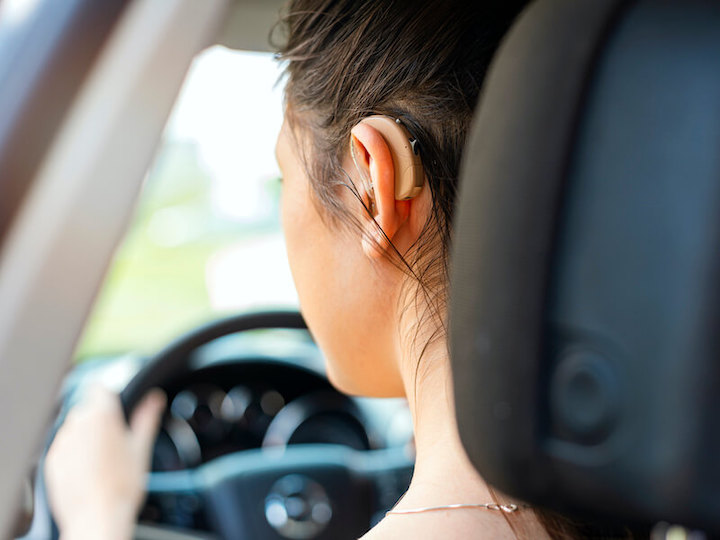 Back-side view of a woman wearing a hearing aid as she's grabbing the steer to drive a car.
