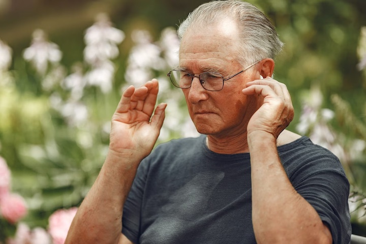 An elderly man in glasses putting on hearing aids.