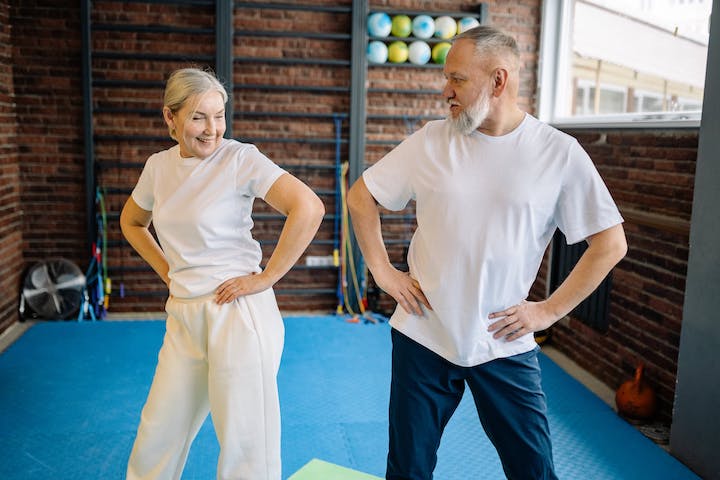 An elderly pair swaying their hips with smiles on their faces in midst of an exercise session.