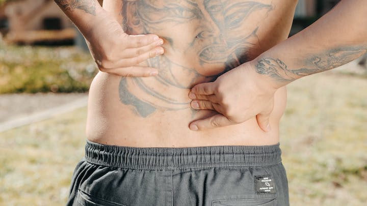 A lower back view of a shirtless man with tattoos pressing on a pain in his lower back.