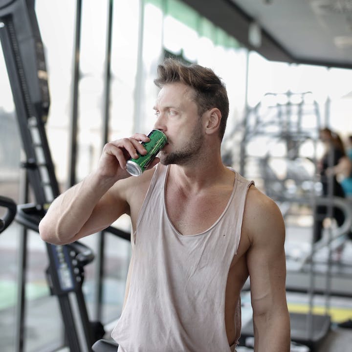 Tired young male athlete drinking energy drink while resting after workout in gym.
