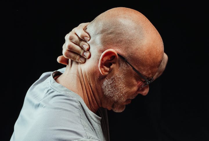 Side view of a middle-aged to-older male in glasses grasping his own neck from behind in pain.