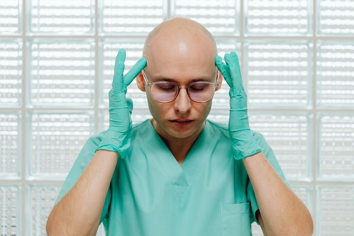A doctor in green medical uniform and green gloves touching two temples of his head with closed eyes and glasses on.