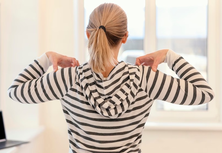 Back view of a woman in a striped hoodie massaging her shoulders to correct her posture in the middle of work.