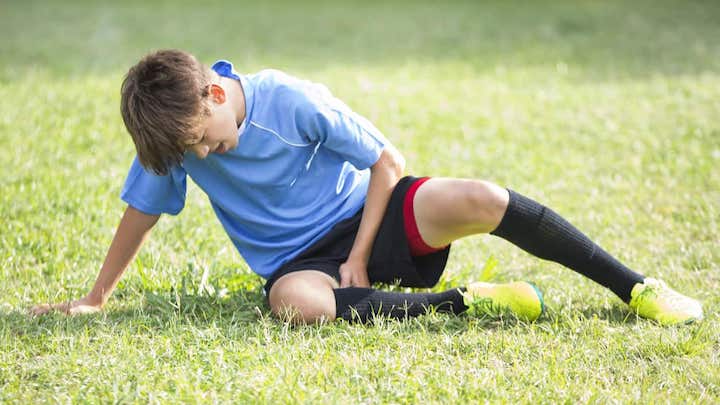 Caucasian teenage boy holds his leg while grimmacing in pain after sustaining injury during soccer game. He is alone on the playing field. He is sitting on the grass. He is wearing a blue jersey, black shorts, black socks and cleats.
