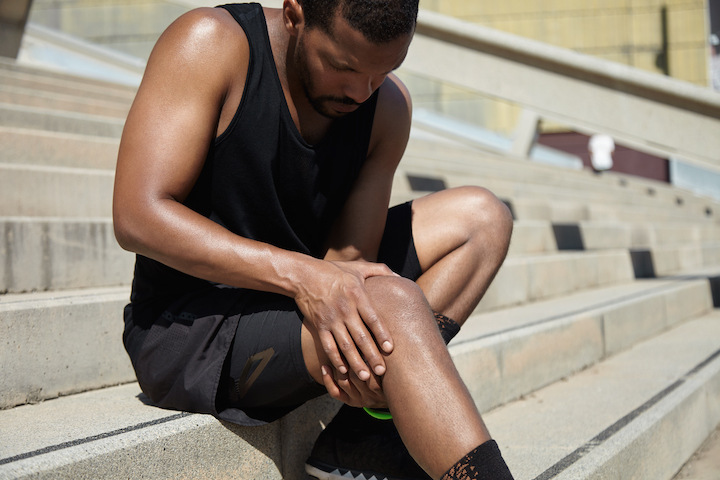 An African-American man clutching his knee in the patellofemoral area in pain while sitting on stairs.
