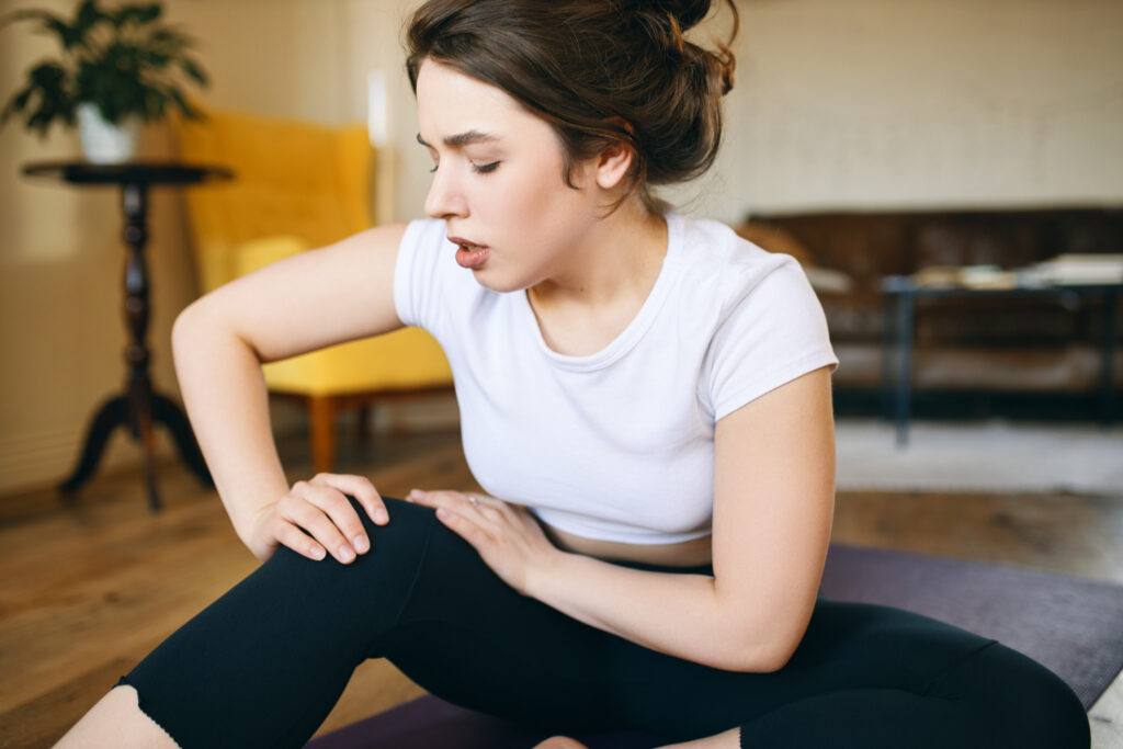 A woman sits on the floor, holding her knee in discomfort, highlighting the struggle with hip bursitis pain.
