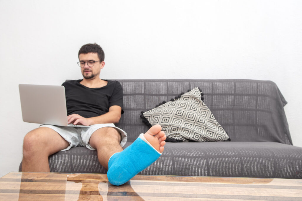 A man with a foot cast sits on a couch, using a laptop to book online physiotherapy sessions for recovery.
