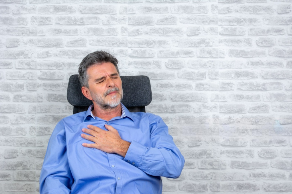  Man sitting in a chair, holding his chest in pain, highlighting the effects of poor posture on health.