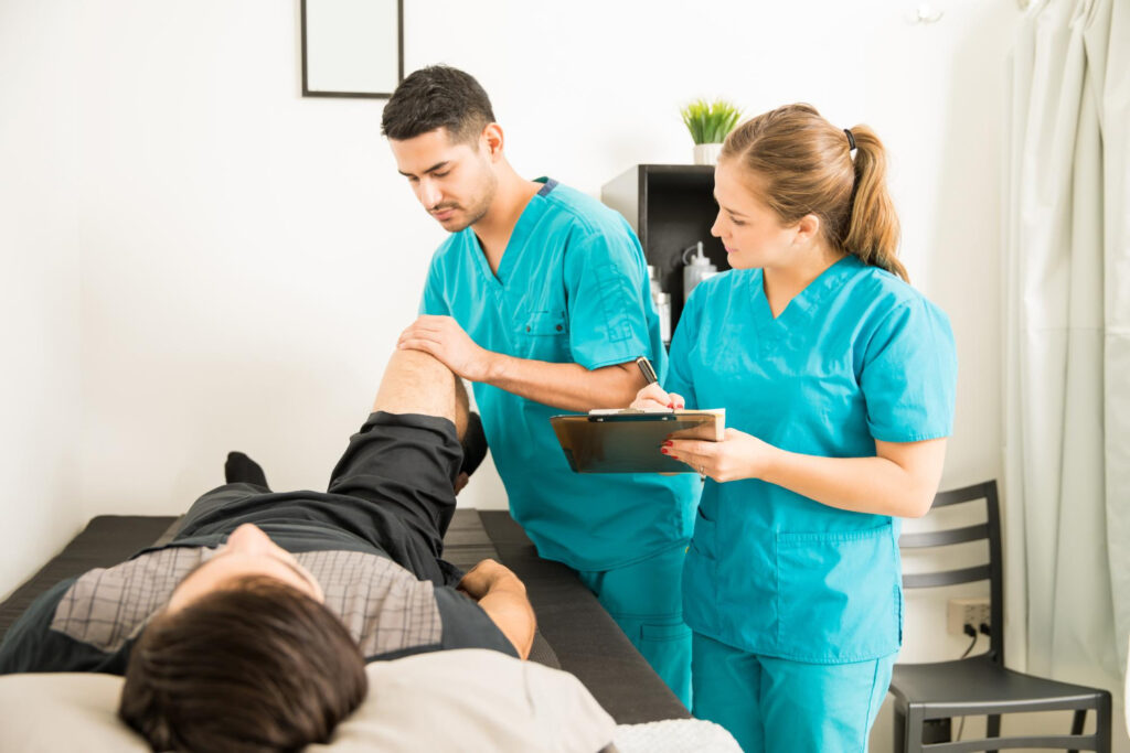 A man rests on a bed while a nurse examines his knee for signs of a calf strain.
