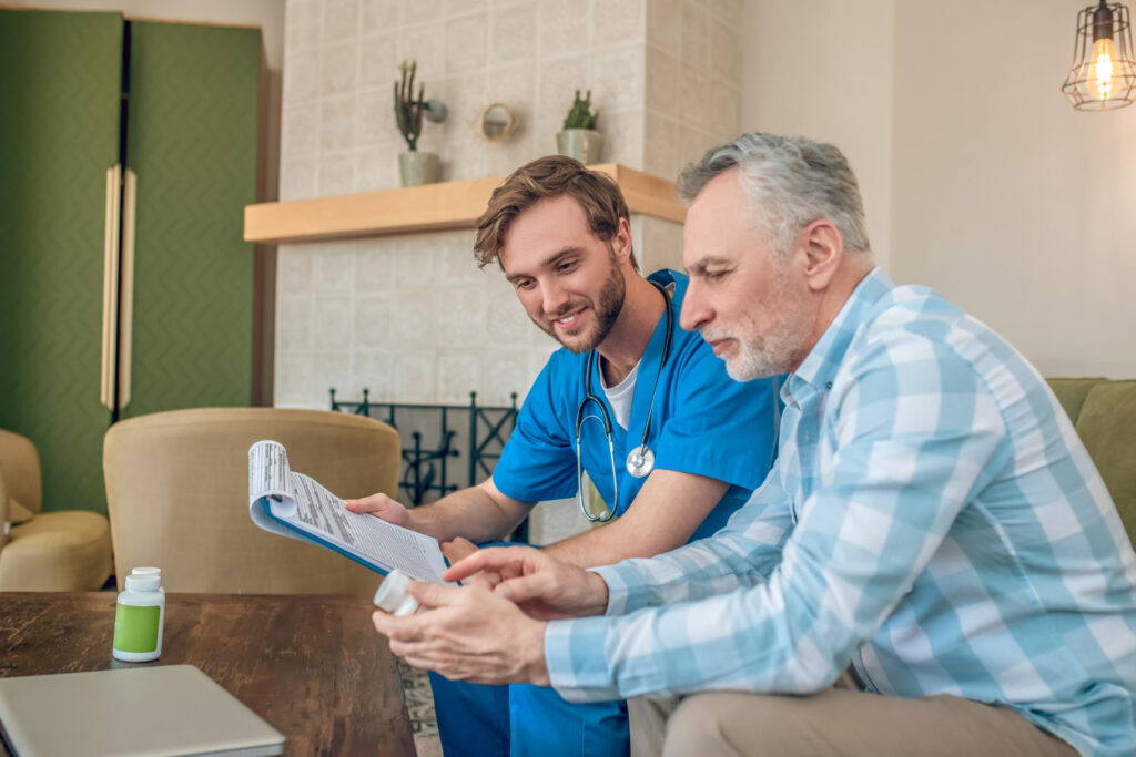 A man and a nurse sit on a couch, reviewing a document about important considerations for home-based rehab.
