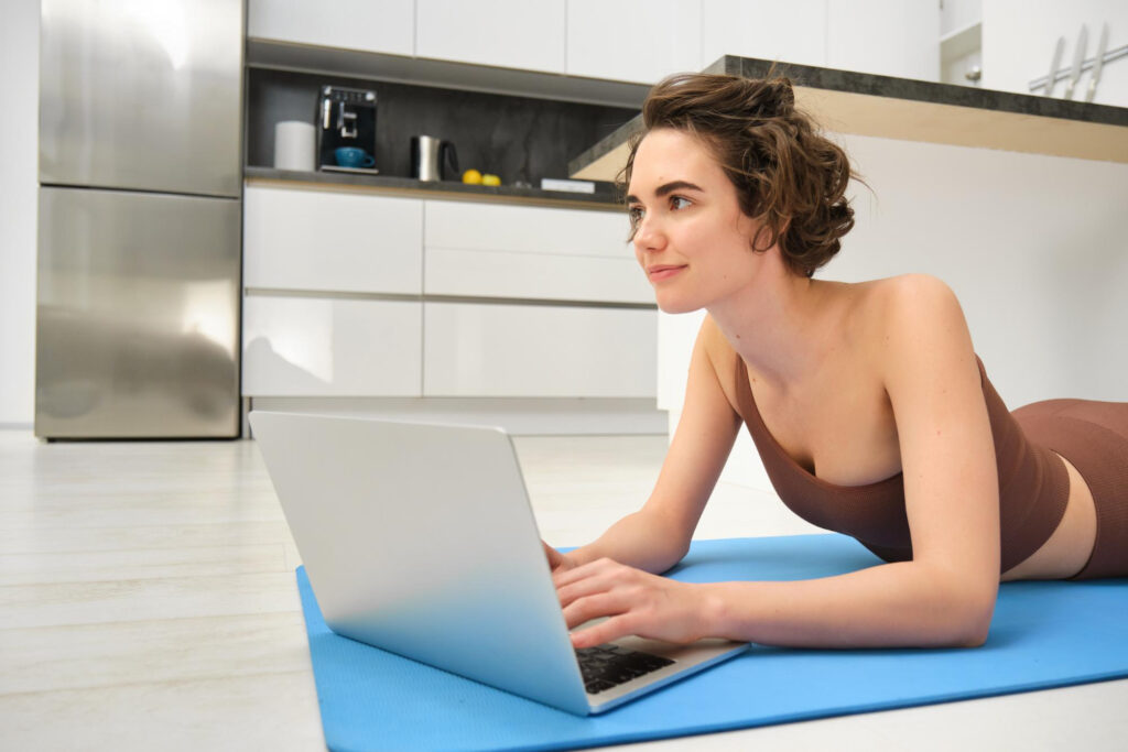 A woman in a bra top lies on a yoga mat, engaging with her laptop for online physiotherapy in Australia.