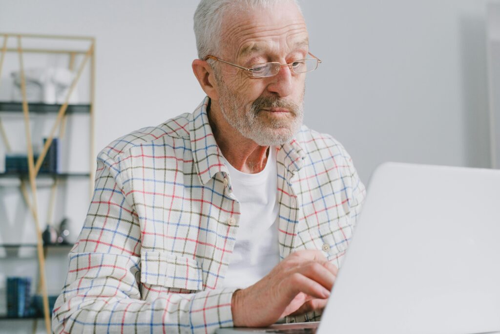 An older man in glasses and a plaid shirt navigates challenges while using a laptop.