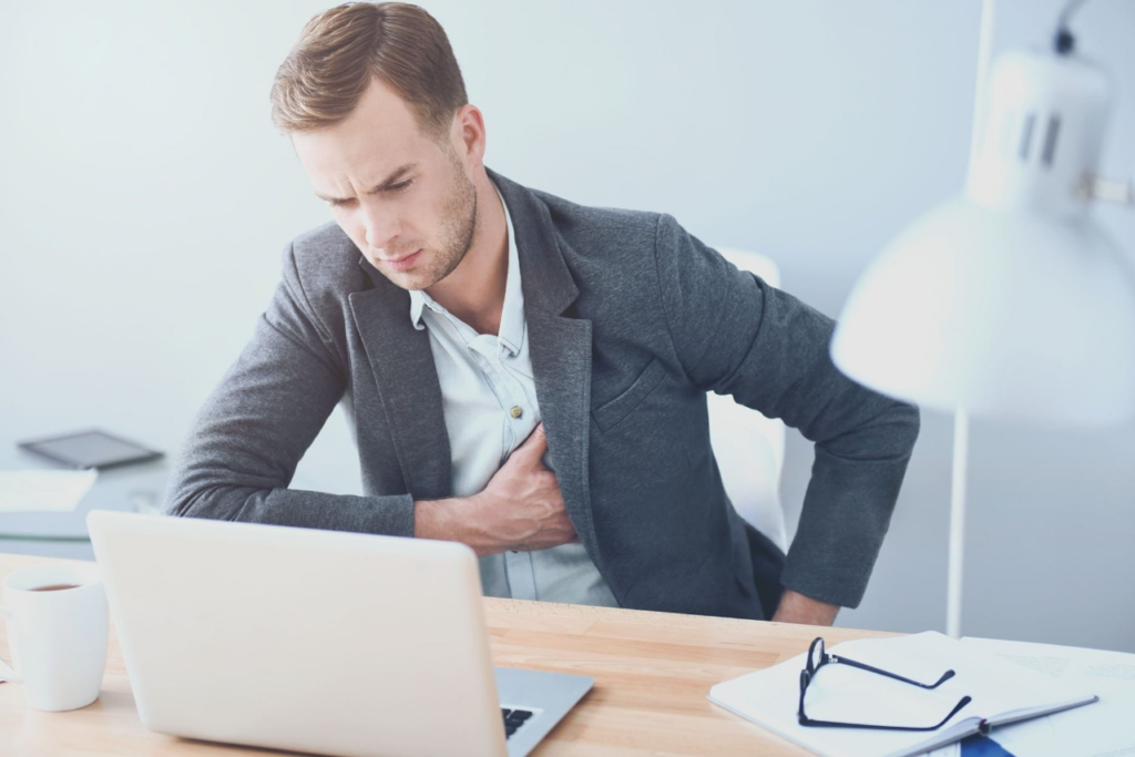 A man at a desk, hands on his chest, showing signs of discomfort from poor posture and chest pain symptoms.