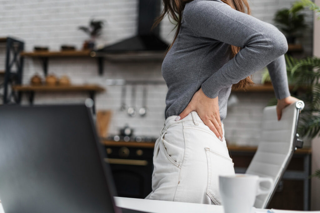 A woman sitting at a laptop, visibly uncomfortable, holding her lower back due to pain while working.