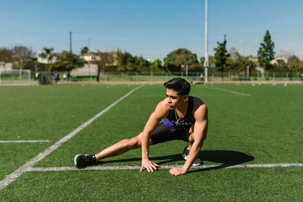 A man kneels on the grass of a soccer field, demonstrating a stretch to loosen tight leg muscles for improved performance.