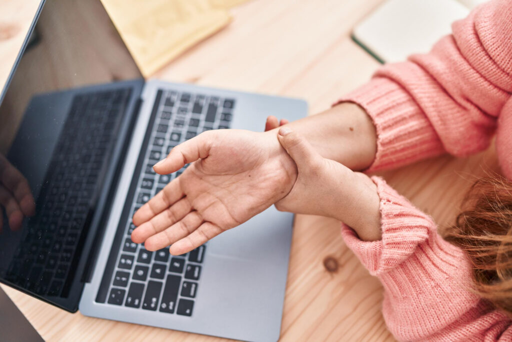 A woman sitting at a laptop, holding her wrist, possibly indicating discomfort or weakness in her wrist.