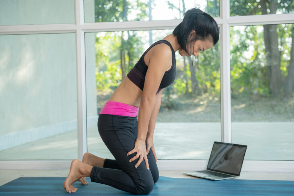 A woman in a sports bra and pink shorts kneels on a yoga mat, focusing on stretching tight bum muscles.