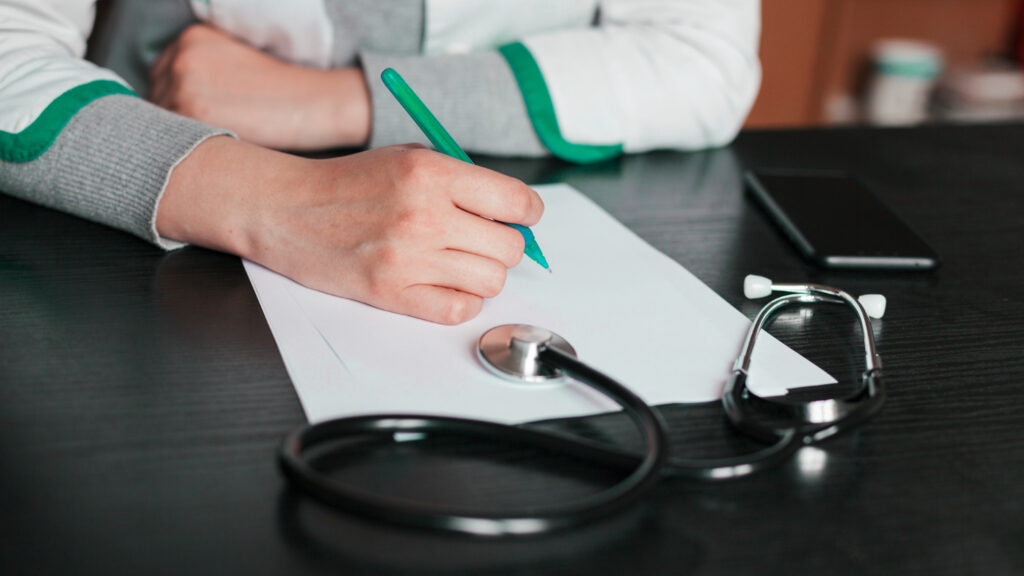 A person using a stethoscope while writing notes on paper, symbolizing diagnosis and assessment in healthcare.