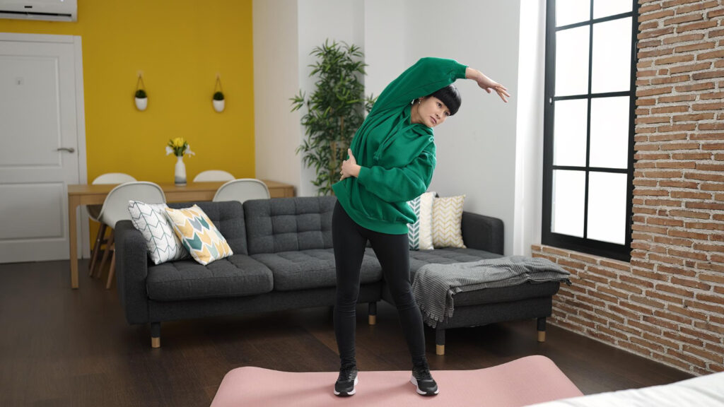 A woman in a green sweatshirt practices yoga in her cozy living room, focusing on preventing floating rib pain.
