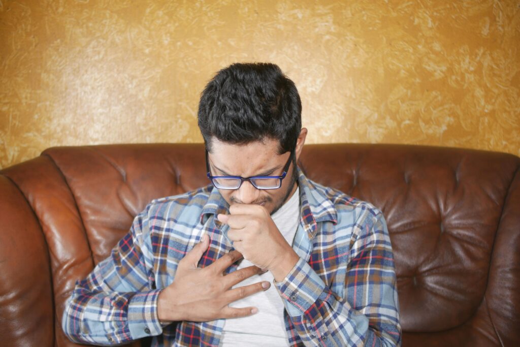 A man with glasses confidently sits on a couch, showcasing his chest, while discussing tips to prevent rib pain from coughing.