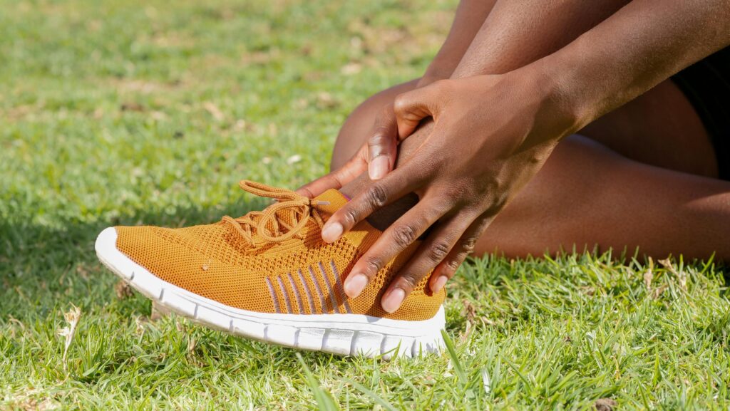 A woman grips her shoe on the grass, suggesting she may be dealing with ankle pain while flexing her foot.