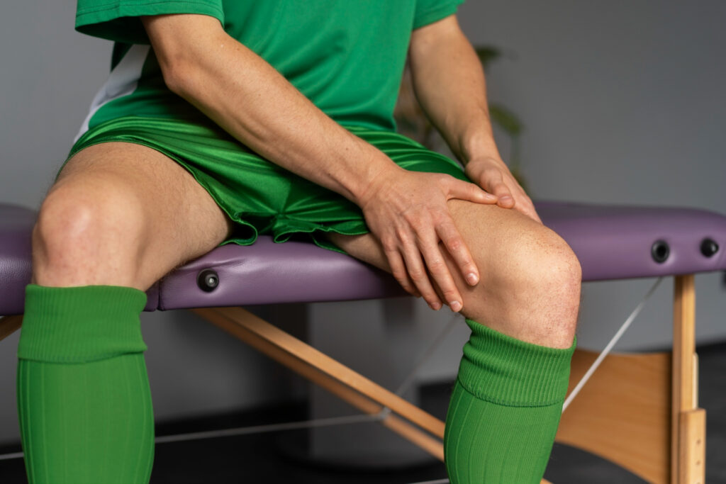 A man seated at a table, visibly distressed, holding his knee, indicating pain associated with shin splints.